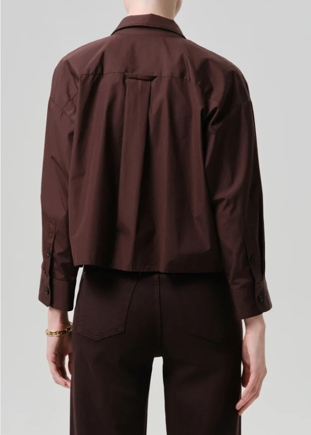 A model shown from the back wearing a dark brown cropped button-up shirt and matching dark brown trousers against a light gray studio background.