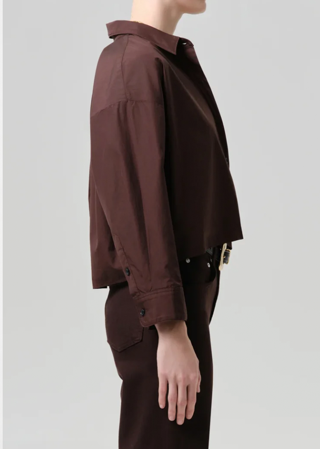 A side-profile view of a model wearing a dark brown cropped button-up shirt and matching dark brown trousers against a light gray studio background.