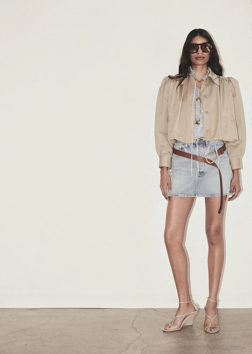 Woman wearing a beige jacket, denim shorts, and sunglasses against a white background