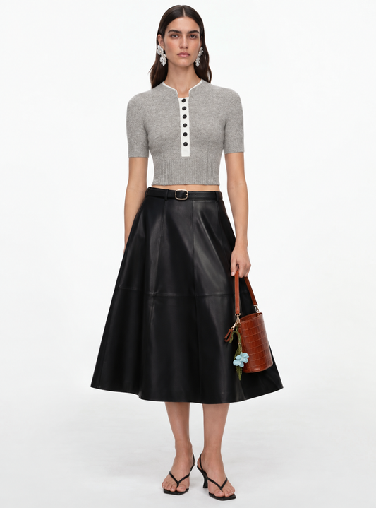 A model stands against a white studio background wearing a gray short-sleeve cropped henley top with a white placket and black buttons, paired with a black pleated midi skirt, a brown handbag, and black sandals.